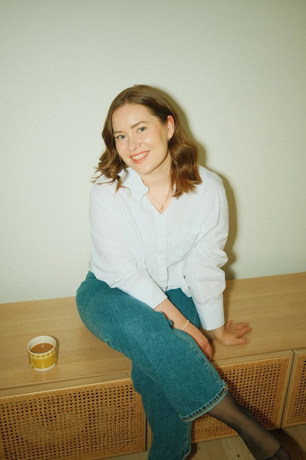 Kuvassa nainen, joka istuu TV-tasolla ja katsoo hymyillen kameraan. Hänen vierellään tason päällä on kahvikuppi.